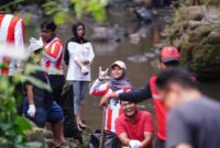 Di tengah rimbunnya tepian sungai, para pemuda yang mengenakan pakaian bernuansa merah-putih dan atribut relawan tampak melempar senyum serta pose penuh semangat ke arah kamera.